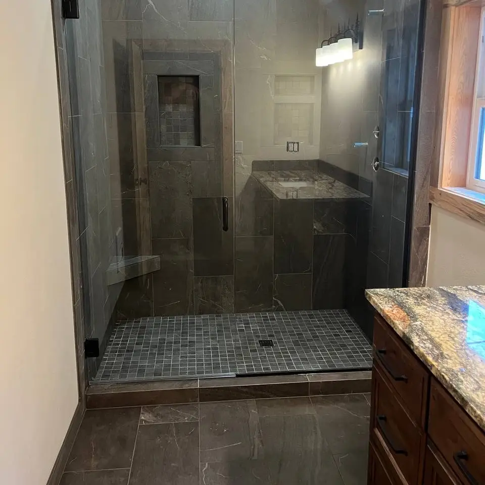 Walk-in shower in charcoal veined tile behind frameless glass, with a mosaic-backed niche, a corner ledge and a small square mosaic floor