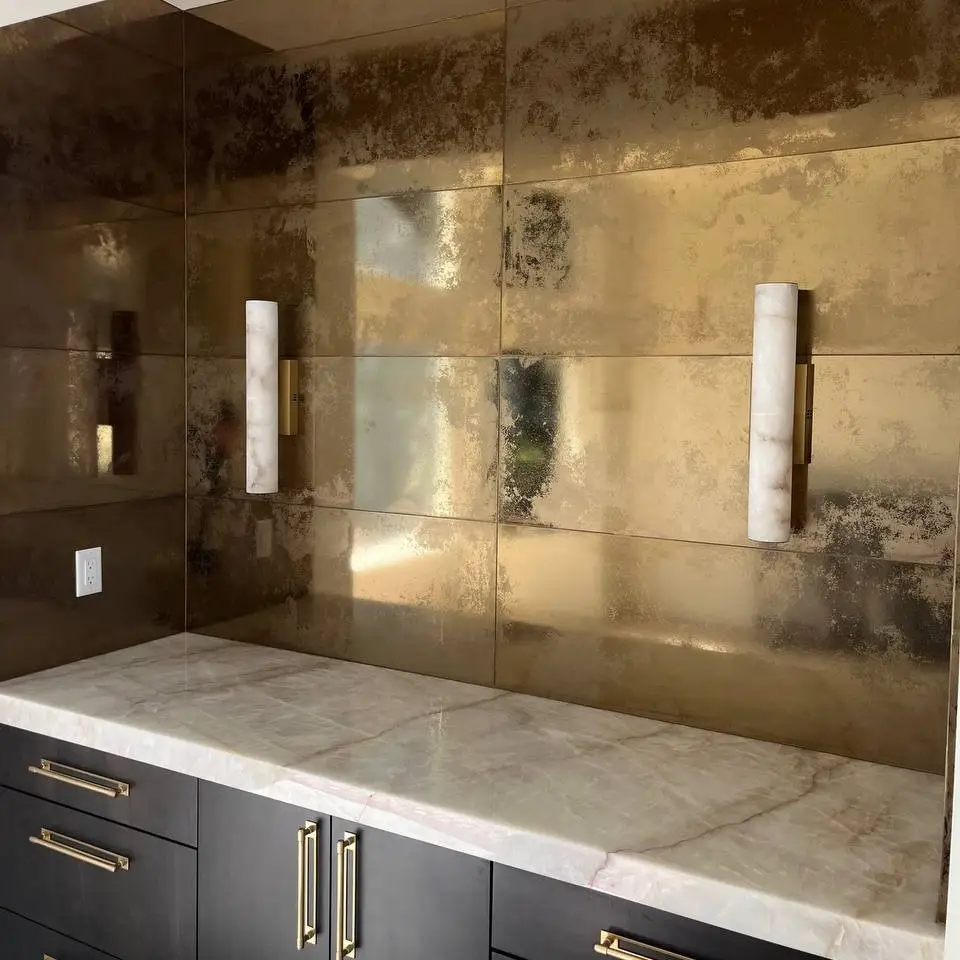 Antiqued gold metallic tile backsplash behind two wall sconces above a white stone counter