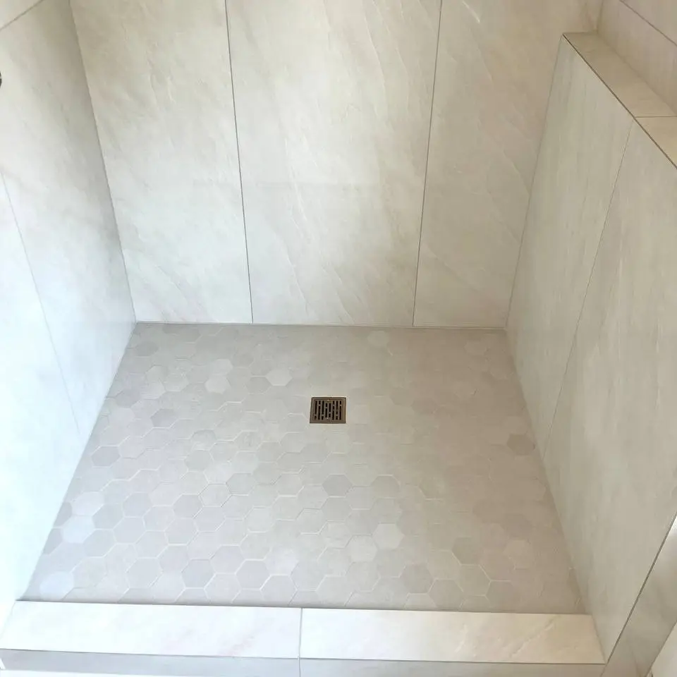 Walk-in shower in large-format light marble-look tile with a hexagon mosaic floor, a square drain and a tile-capped knee wall