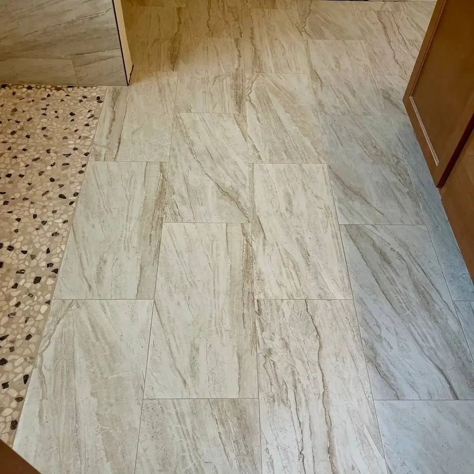 Large beige marble-look floor tile laid in an offset pattern, meeting a pebble mosaic shower floor