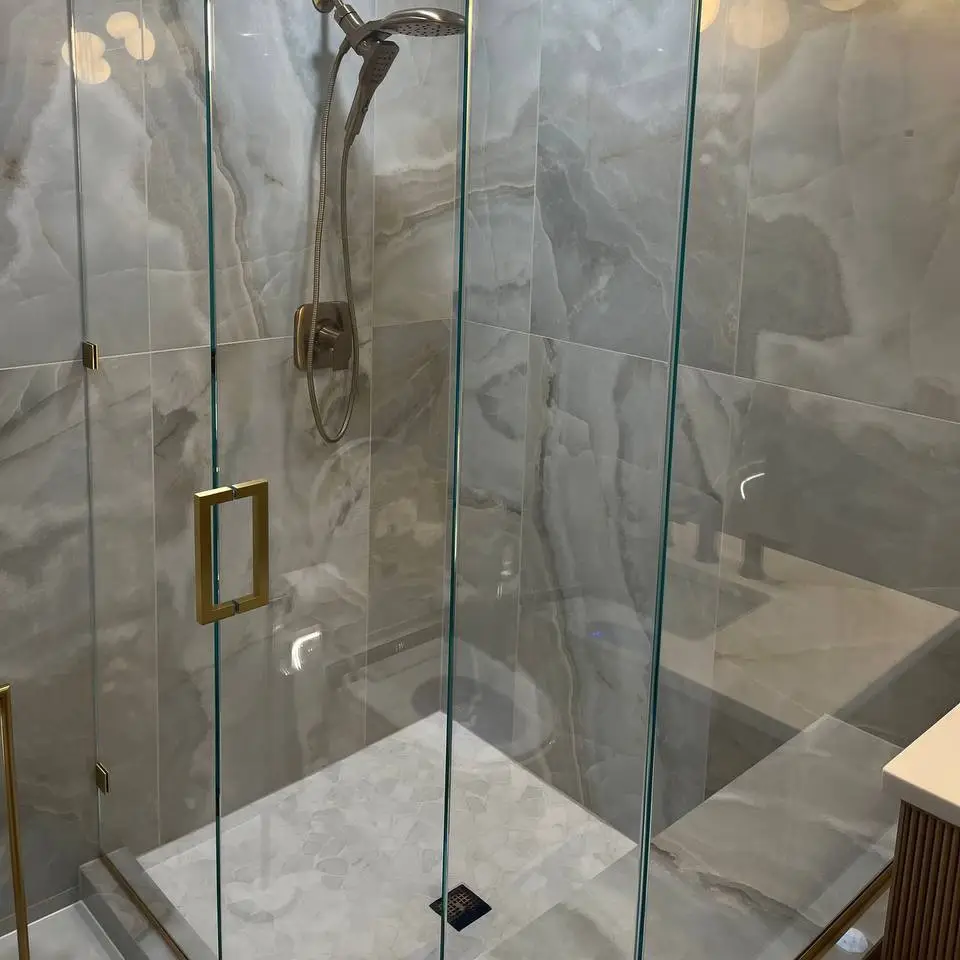 Frameless glass shower lined in large onyx-look tile, with a brushed-gold handheld shower and a white stone mosaic floor