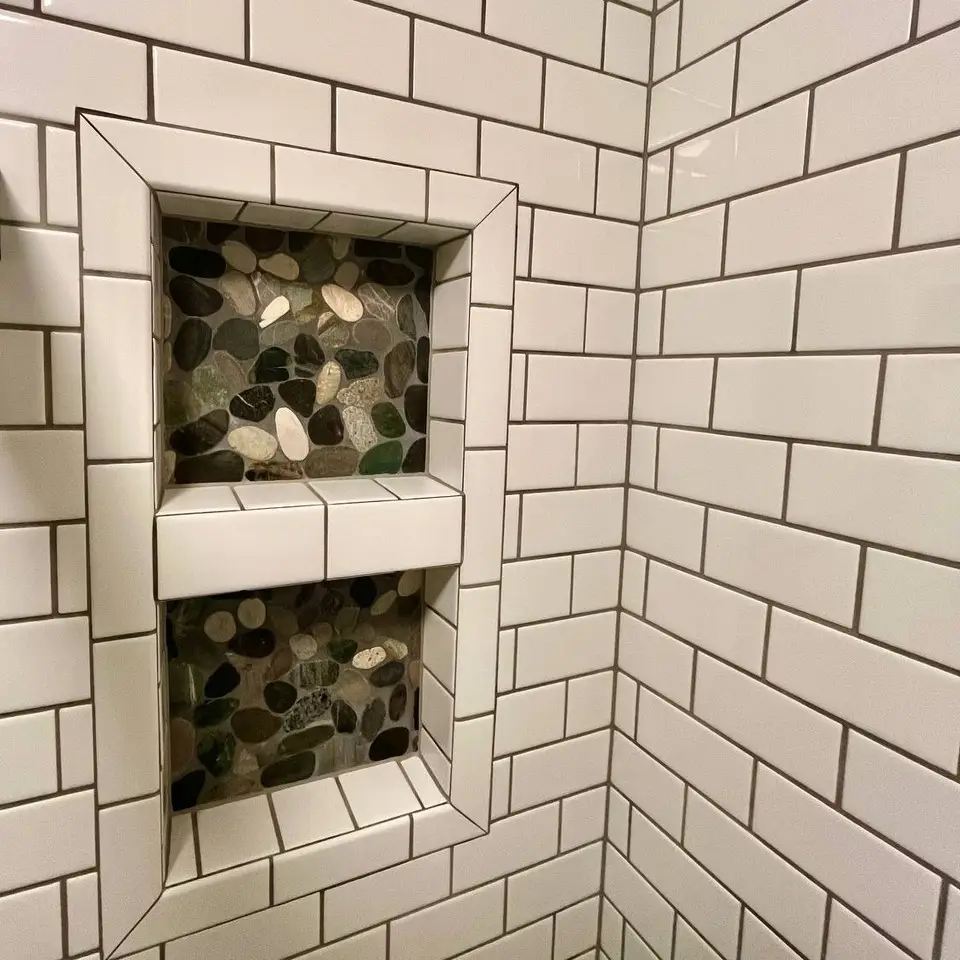 Double shower niche framed in white subway tile with dark grout, backed with river-pebble mosaic
