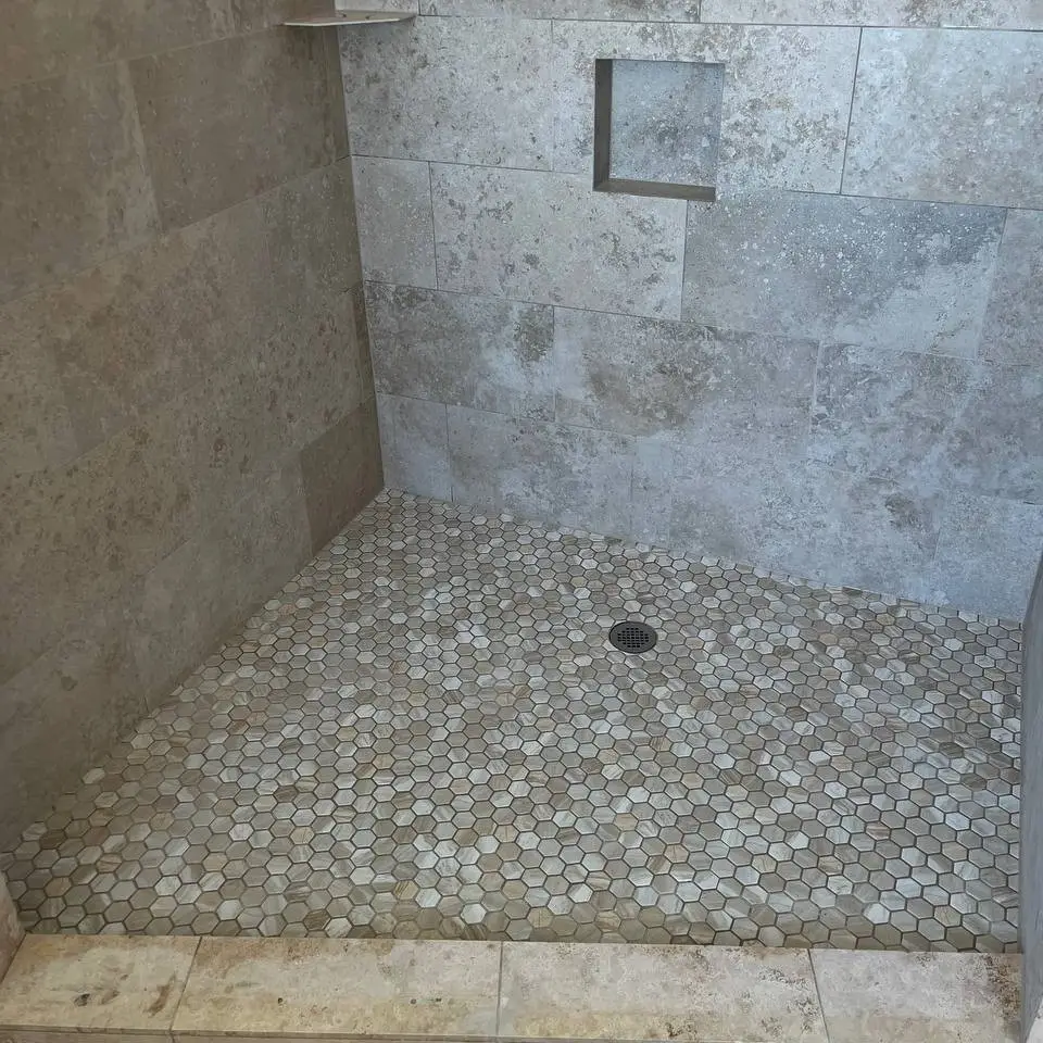 Walk-in shower in grey stone-look wall tile with a recessed niche, above a hexagon stone mosaic floor and a tiled curb