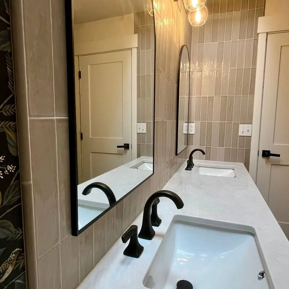 Glossy taupe vertical tile behind a double vanity, with the outlets cut cleanly into the tile beside black-framed mirrors